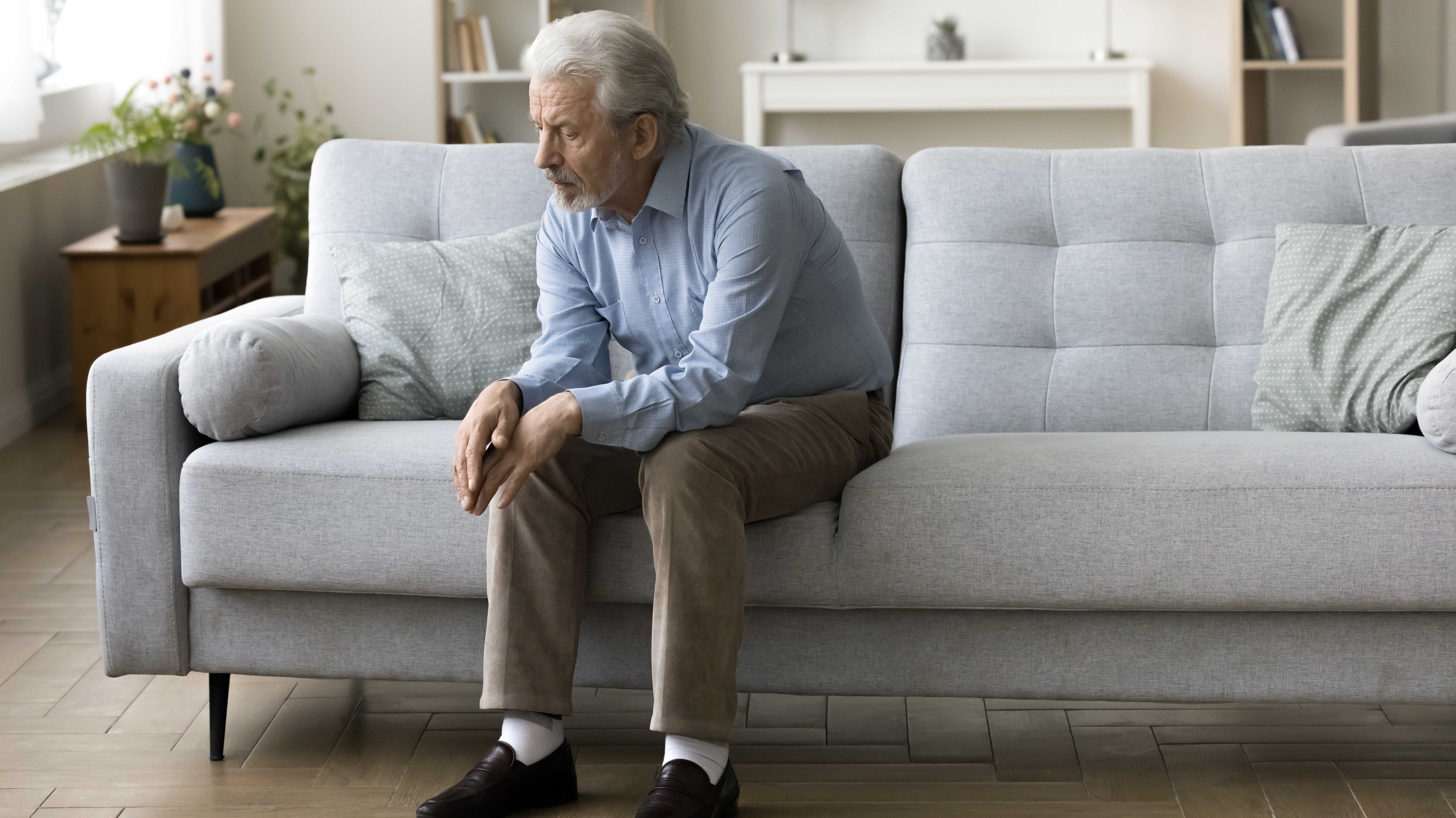Trauriger, einsamer, alter, grauer Haar, allein auf dem Sofa zu Hause sitzender Mann, der schlecht darüber nachgedacht wurde, unter Depressionen leidet, Apathie leidet, mit Verlust fertig ist, schlechte Nachrichten über Gesundheitsversorgung, Familienprob